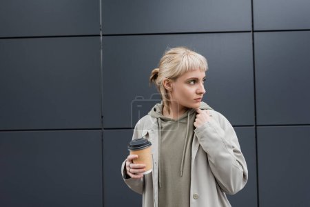 Photo pour Jeune femme à la mode avec les cheveux blonds avec frange debout dans le manteau et sweat à capuche tout en tenant tasse en papier avec café à emporter près du bâtiment moderne gris dans la rue, à l'extérieur, la vie urbaine - image libre de droit