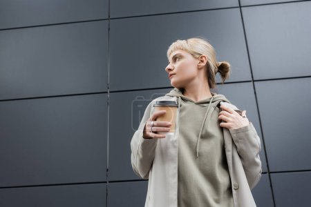 stylish young woman with blonde hair with bangs looking away and standing in coat and hoodie while holding paper cup with takeaway coffee near grey modern building on street, outside, urban living 