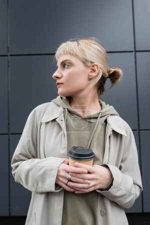 Photo for Stylish young woman with blonde hair with bangs standing in coat and hoodie while holding paper cup with takeaway coffee near grey modern building on street, outside, urban living, look away - Royalty Free Image