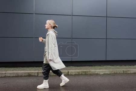side view of young woman with blonde hair with bangs walking in coat, black leather pants, hoodie and boots while holding paper cup with takeaway coffee near grey modern building on street 