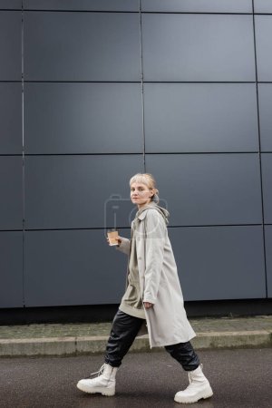Photo for Full length of trendy woman with blonde hair with bangs walking in coat, black leather pants, hoodie and boots while holding paper cup with takeaway coffee near grey modern building on street - Royalty Free Image