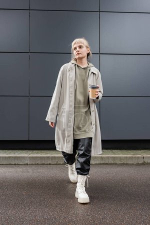 full length of stylish young woman with blonde hair with bangs walking in coat, black leather pants, hoodie and boots while holding paper cup with takeaway coffee near grey modern building on street 