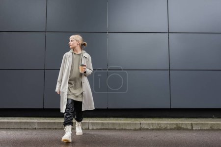 Photo for Full length of young woman with blonde hair with bangs walking in coat, black leather pants, hoodie and boots while holding paper cup with takeaway coffee near grey modern building on street - Royalty Free Image