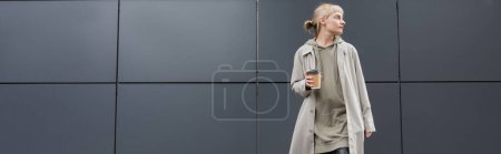 stylische junge Frau mit Pony in Mantel und Kapuzenpulli, während sie Pappbecher mit Kaffee zum Mitnehmen in der Nähe eines grauen modernen Gebäudes auf der Straße hält, draußen, urbanes Leben, wegschauen, Transparent