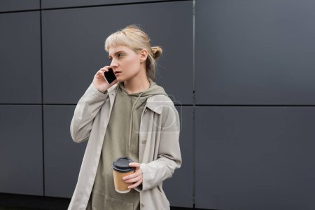 stylish young woman with bangs and blonde hair holding paper cup with coffee to go while talking on smartphone and standing in hoodie and coat near grey modern building on urban street 