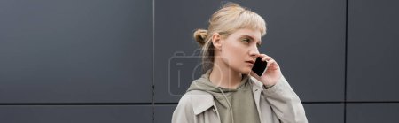 Photo for Stylish young woman with bangs and blonde hair talking on smartphone and standing in hoodie and coat near grey modern building on urban street, banner, looking away, urban lifestyle - Royalty Free Image