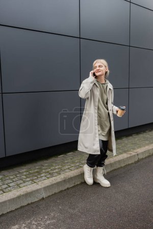 glückliche junge Frau mit Pony und blonden Haaren, die Pappbecher mit Coffee to go hält, während sie mit dem Smartphone spricht und in Kapuzenpulli und Mantel in der Nähe eines grauen modernen Gebäudes an der Stadtstraße steht 