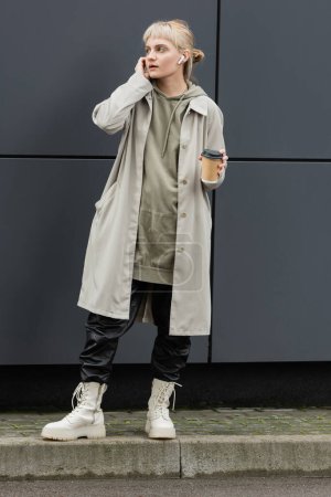mujer joven de moda con flequillo sosteniendo taza de papel con café para llevar mientras se ajustan los auriculares inalámbricos y de pie en traje de moda, sudadera con capucha y abrigo cerca de edificio moderno gris en la calle urbana 
