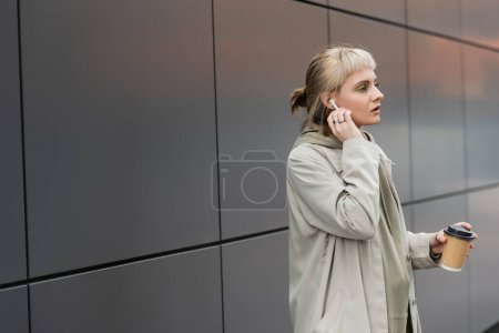Photo pour Femme blonde avec frange tenant tasse en papier avec café à emporter tout en ajustant les écouteurs sans fil et debout dans une tenue à la mode, près du bâtiment moderne gris sur la rue urbaine, détourner les yeux - image libre de droit