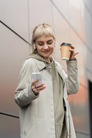 glückliche junge Frau mit Pony hält Pappbecher mit Coffee to go in der Hand und steht im trendigen Outfit und mit drahtlosen Kopfhörern in der Nähe eines grauen modernen Gebäudes an der Stadtstraße 