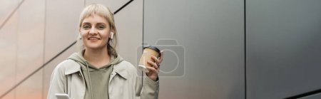sonriente mujer joven con flequillo sosteniendo taza de papel con café para ir y de pie en traje de moda y auriculares inalámbricos mientras se utiliza el teléfono inteligente cerca de edificio moderno gris en la calle urbana, pancarta