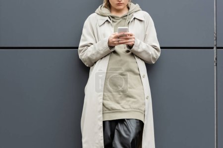 Ausgeschnittene Ansicht einer modischen jungen Frau, die in trendiger Kleidung in der Nähe einer grauen Hauswand an der Stadtstraße steht und SMS schreibt, Mantel mit Kapuzenpulli, Teilansicht