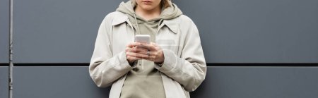 Photo for Cropped view of fashionable young woman standing in trendy clothes and texting on smartphone near grey wall of modern building on urban street, coat with hoodie, partial view, banner - Royalty Free Image