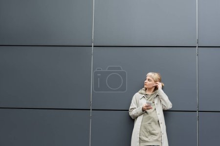 jeune femme à la mode avec une frange debout dans des vêtements à la mode et des écouteurs sans fil tout en utilisant un smartphone près du bâtiment moderne gris dans la rue urbaine, manteau avec capuche, détournant les yeux