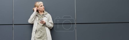 Photo for Stylish young woman with bangs standing in trendy clothes and wireless earphones while using smartphone near grey modern building on urban street, coat with hoodie, looking away, banner - Royalty Free Image