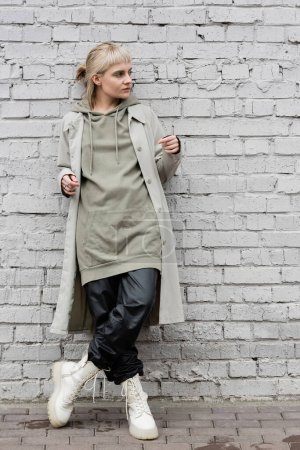 Photo for Fashonable young woman with makeup, blonde hair, bangs, in stylish outfit, long hoodie, coat, black leather pants and beige boots standing near grey brick wall and looking away - Royalty Free Image