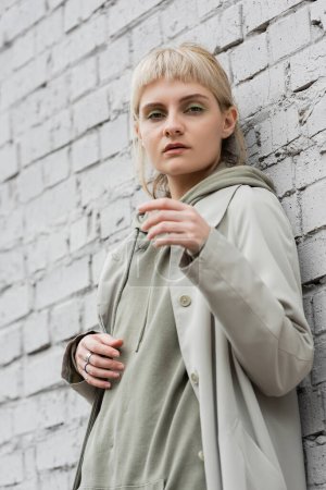 Photo for Young woman with makeup, blonde hair, bangs, in stylish outfit standing near grey brick wall of modern building and looking at camera while posing outdoor, coat, fashion trend - Royalty Free Image
