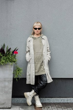 full length of young woman in stylish sunglasses, long hoodie, coat, black leather pants and beige boots standing near grey wall of modern building and posing with hands in pockets near green plant 