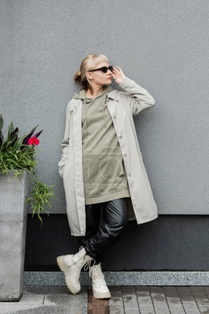 full length of young woman in stylish sunglasses, long hoodie, coat, black leather pants and beige boots standing near grey wall of modern building and posing with hand in pocket near green plant 