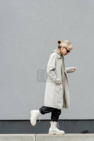 longitud completa de la mujer joven en gafas de sol con estilo, sudadera con capucha larga, abrigo, pantalones de cuero negro y botas beige caminando con la mano en el bolsillo cerca de la pared gris del edificio moderno y mirando hacia abajo 