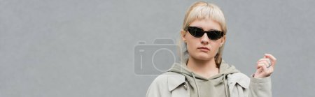 stylish and young woman with bangs and blonde hair standing in trendy sunglasses and comfortable clothes while looking at camera isolated on grey background in studio, hoodie, banner 