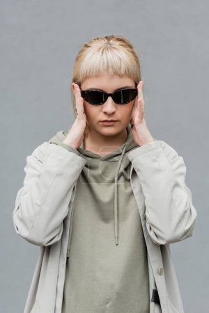 stylish and young woman with bangs and blonde hair standing in trendy sunglasses and comfortable clothes while looking at camera isolated on grey background in studio, hoodie 