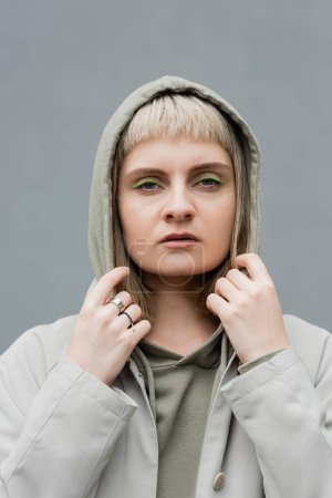 Photo for Stylish and young woman with bangs and blonde hair standing with hood on head and comfortable clothes while looking at camera isolated on grey background in studio, hoodie - Royalty Free Image