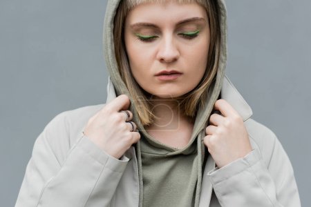 stylish and young woman with bangs, green eye shadows and blonde hair standing with hood on head and comfortable clothes while looking down isolated on grey background in studio, hoodie 
