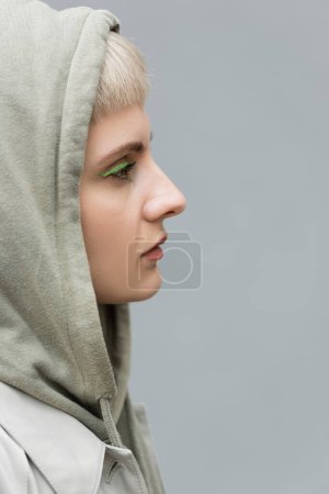 Photo for Side view of stylish and young woman with bangs, green eye shadows and blonde hair standing with hood on head and comfortable clothes while looking away isolated on grey background in studio, hoodie - Royalty Free Image