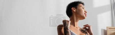 young and sexy tattooed woman with short brunette hair touching straps of silk bra while looking away and dreaming near white wall in light bedroom at home, banner