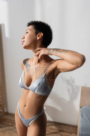 young and irresistible woman with sexy tattooed body and short brunette hair touching neck and looking away while standing in grey silk bra and panties near white wall in light bedroom at home