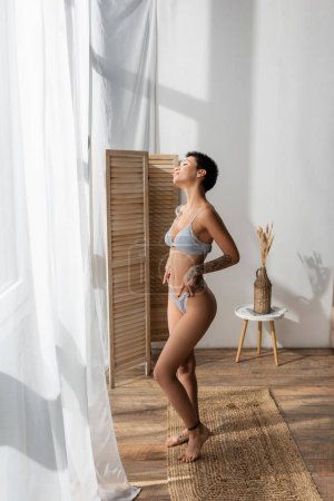 Photo for Full length of stunning woman with sexy tattooed body enjoying natural light while standing on wicker rug near white curtain, room divider and bedside table with vase and spikelets in modern bedroom - Royalty Free Image
