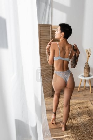 back view of sultry woman with sexy tattooed body wearing lace panties and bra, standing on wicker rug, looking away near white curtain, room divider and vase with spikelets on bedside table 