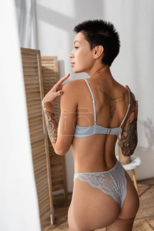 Photo for Charming woman with short brunette hair and sexy tattooed body, wearing lace panties and bra, touching shoulders while looking away near room divider in modern bedroom - Royalty Free Image