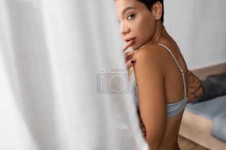junge, sexy und faszinierende Frau in BH, die die Lippen berührt und in die Kamera schaut, während sie in der Nähe des Bettes steht und den weißen Vorhang vor verschwommenem Vordergrund im heimischen Schlafzimmer, Boudoir-Fotografie