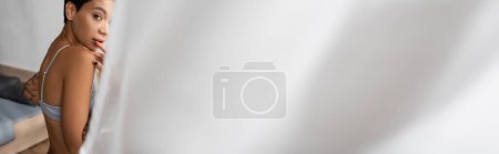 Photo for Young, sexy and flirtatious woman in bra touching lips and looking at camera while standing near bed and white curtain on blurred foreground in bedroom at home, banner - Royalty Free Image