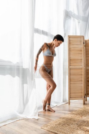 full length of young, tattooed and seductive woman with short brunette hair standing in grey silk lingerie in natural light near white curtain, wicker rug and room divider in modern bedroom at home