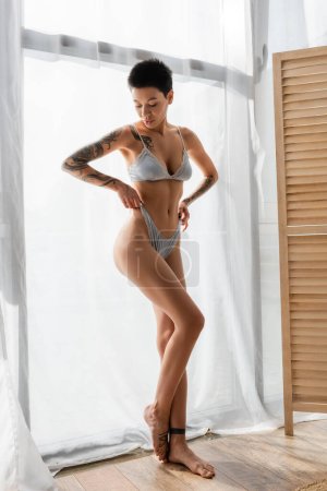 Photo for Full length of young and intriguing woman with short brunette hair and sexy tattooed body pulling grey silk panties while standing near white curtain and room divider in modern bedroom - Royalty Free Image