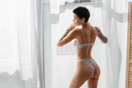 young and stunning woman with sexy tattooed body and short brunette hair wearing lace panties and bra while standing in natural light near white curtain in modern bedroom at home