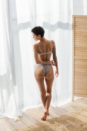 Foto de Longitud completa de mujer joven, tatuada y tentadora con nalgas sexy de pie en luz natural cerca de la cortina blanca, separador de habitaciones y alfombra de mimbre en el dormitorio moderno - Imagen libre de derechos