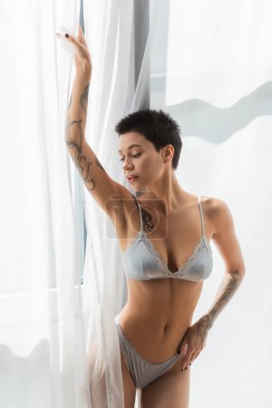 young and tempting woman with short brunette hair, tattooed body and sexy breast standing with grey silk lingerie and posing with raised hand near white curtain in bedroom at home