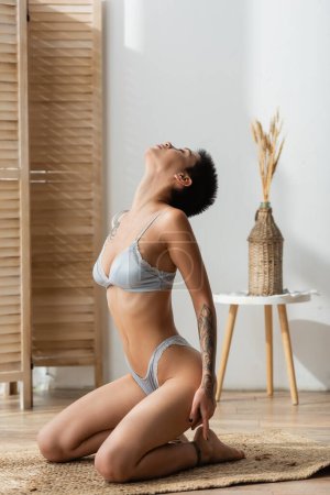 full length of young tattooed and passionate woman in lingerie sitting in seductive pose with thrown back head on wicker rug near room divider, bedside table and vase with spikelets in bedroom