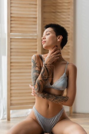 young and tempting woman with tattooed body sitting in seductive pose in grey silk lingerie, touching neck and looking away near room divider on blurred background in bedroom