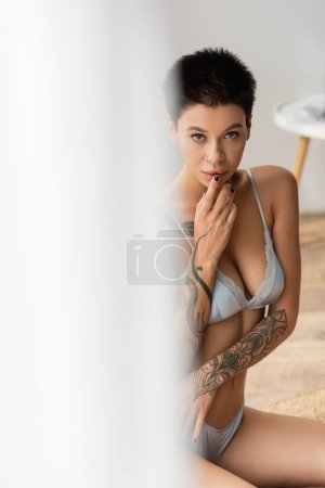 intriguing and seductive tattooed woman with short brunette hair and sexy bust wearing grey silk bra, touching lips and looking at camera in modern bedroom on blurred foreground