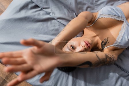 Draufsicht auf charmante und provokante junge Frau mit sexy tätowiertem Körper, die im BH mit erhobenen Händen liegt und auf grauem Bettzeug auf verschwommenem Vordergrund in die Kamera blickt