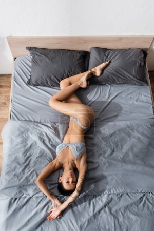 vista superior de la impresionante mujer tatuada con cuerpo sexy y pelo corto morena puesta en ropa de cama gris en lencería de seda y mirando a la cámara, fotografía boudoir, arte de la seducción