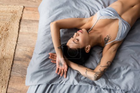 top view of sensual young woman in silk bra, with short brunette hair and sexy tattooed body laying on grey bedding near wicker rug on floor in bedroom at home