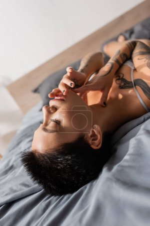 Photo for Young, stunning and sexy woman with closed eyes, tattooed body and short brunette hair touching face while laying on grey bedding on blurred background - Royalty Free Image