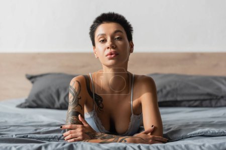 young seductive woman in bra, with short brunette hair and sexy tattooed body looking at camera while laying on grey bedding near pillows on blurred background in bedroom, boudoir photography