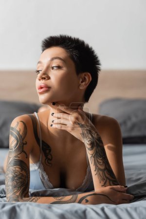 stunning woman with short brunette hair, tattooed body and sexy breast holding hand near neck while laying in bra on grey bedding and looking away on blurred background, boudoir photography